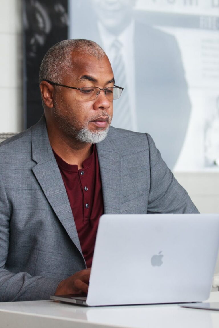 Senior adult man with gray beard working remotely on laptop indoors wearing a blazer.