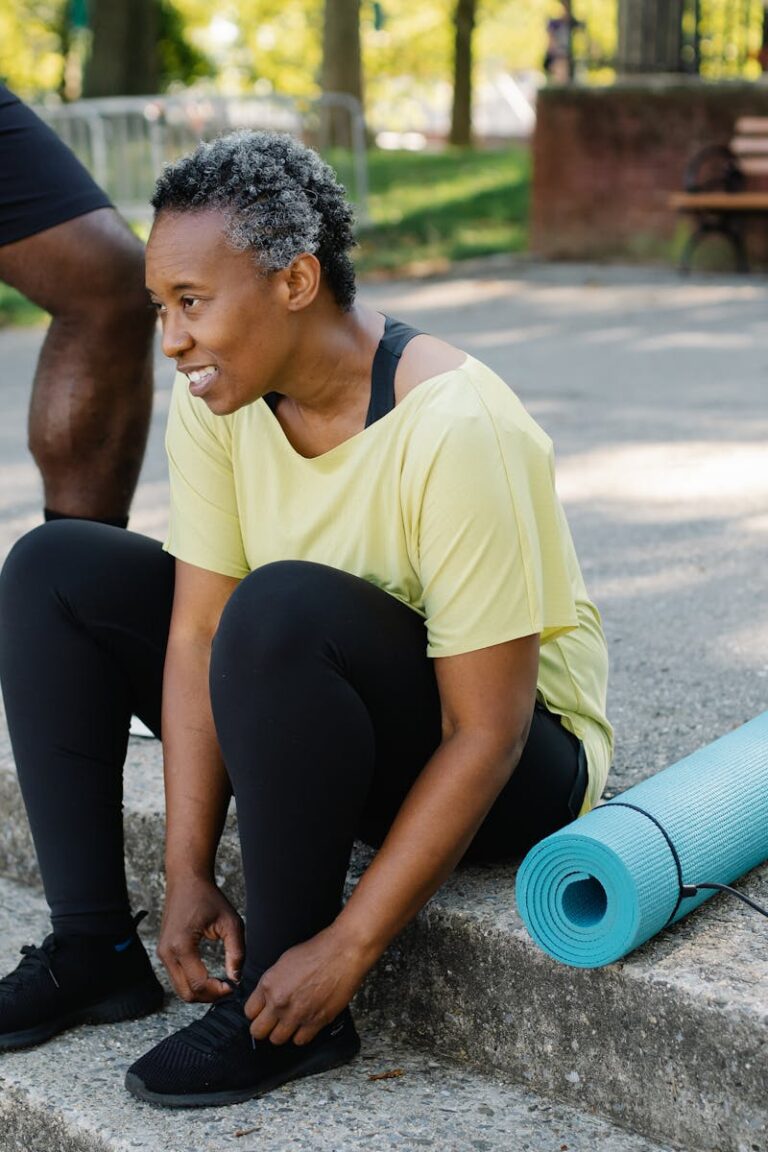 A mature African American woman ties shoelaces beside a yoga mat outdoors.